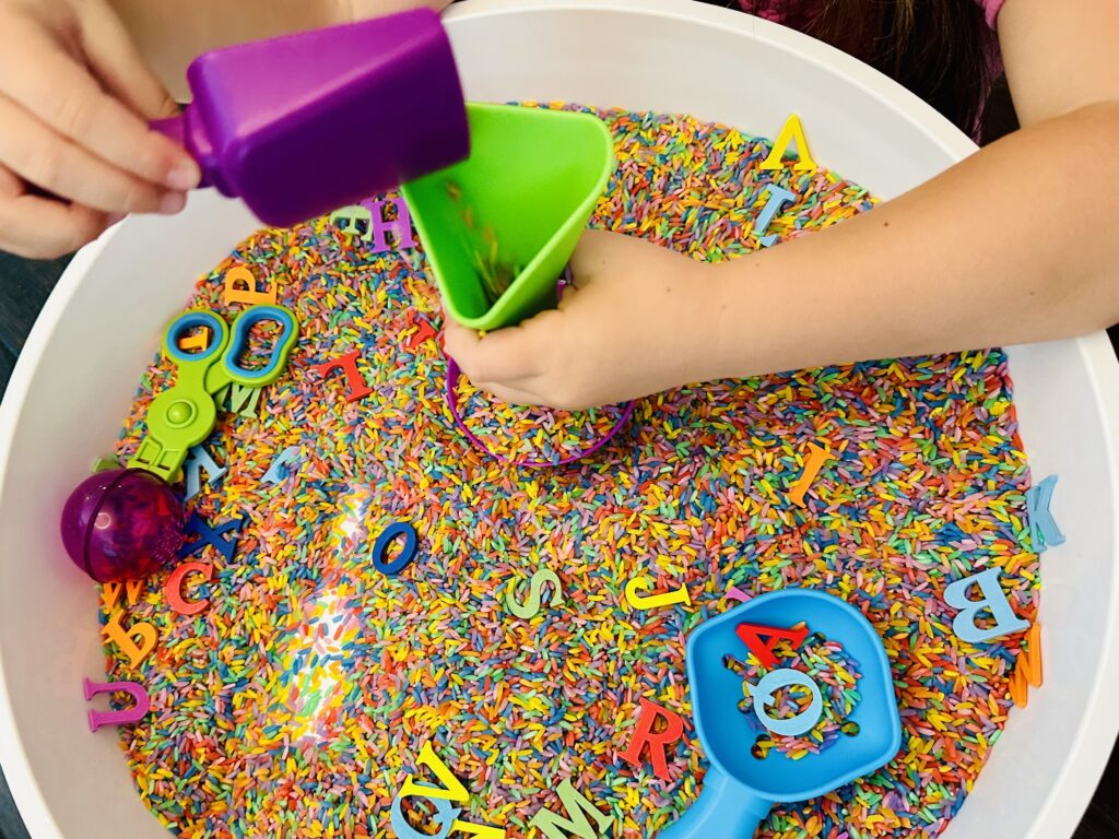 Child using a scoop and funnel in a rainbow rice sensory bin.