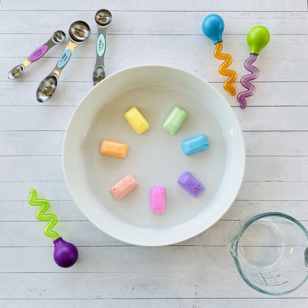 Droppers, measuring spoons, and cup full of vinegar around the frozen fizzy cubes on a baking dish